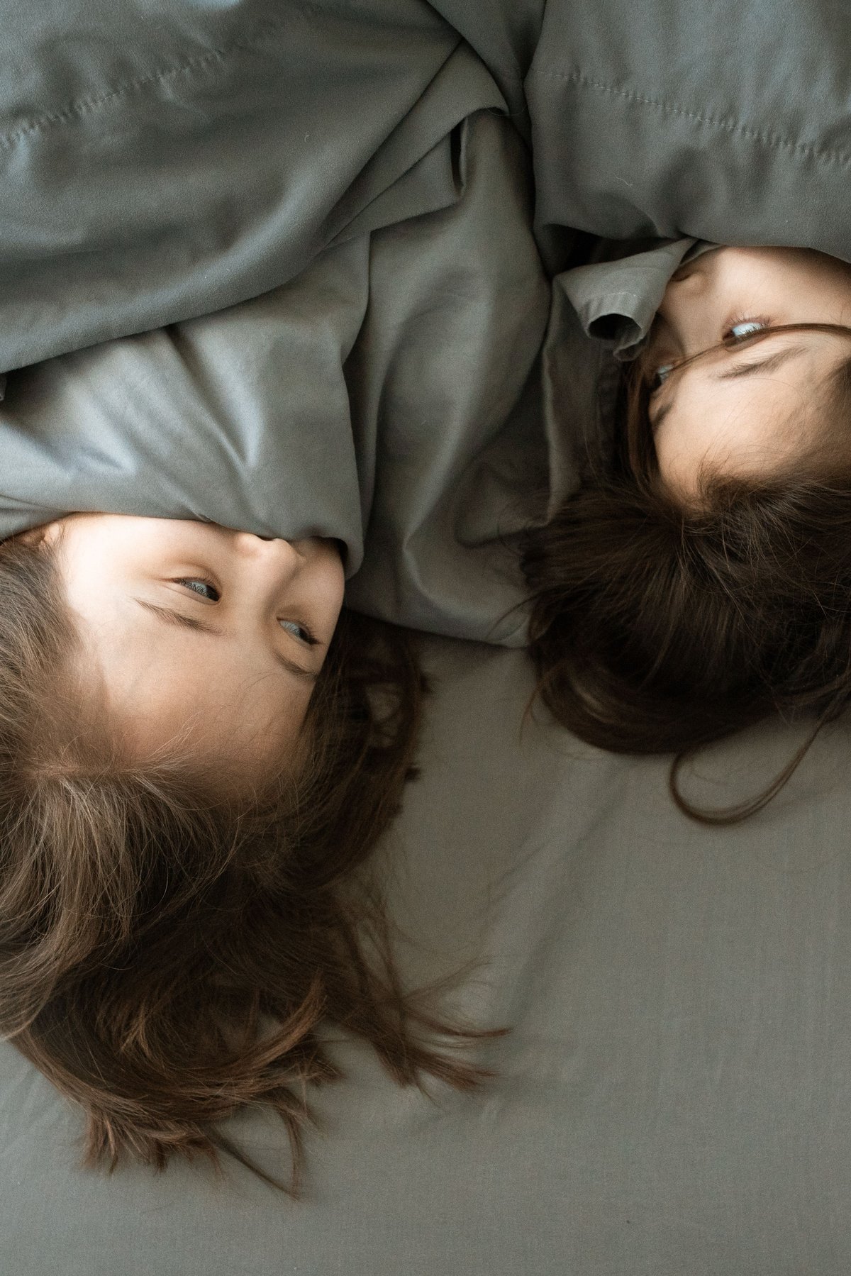 Two children lying in bed together under a grey blanket